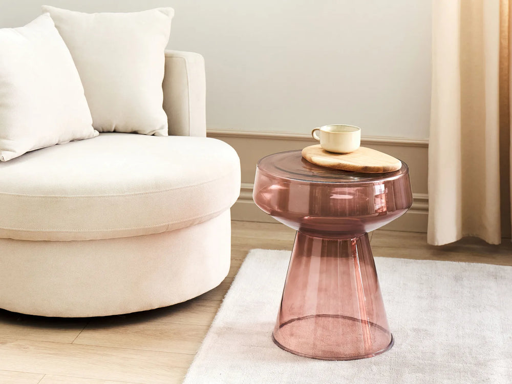 A translucent dusty rose side table with a flared pedestal base, styled in a modern living room next to a cream-colored round sofa.