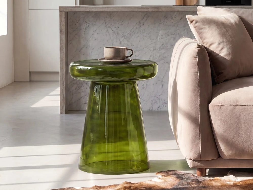 A modern translucent green glass side table with a rounded top and tapered base, styled in a bright living room next to a beige sofa with a cup on top.