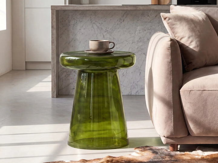 A modern translucent green glass side table with a rounded top and tapered base, styled in a bright living room next to a beige sofa with a cup on top.