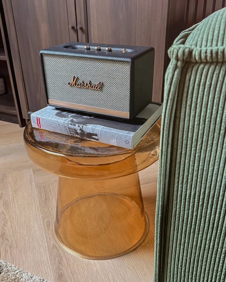 Marshall speaker on a glass side table with a book underneath, in a room setting.