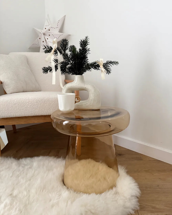 Glass side table with a white vase, cup, and decorative branches in a living room setting.