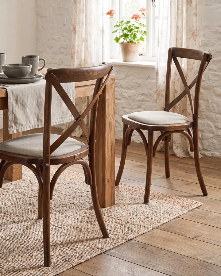Two wooden dining chairs with cream upholstered seats, shown in a bright dining room setting with a table, rug, and window.