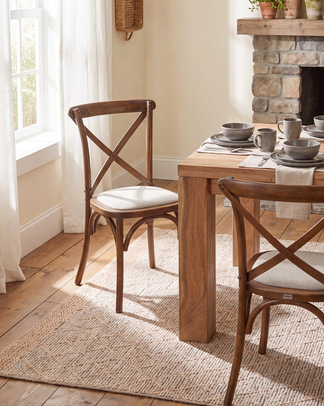A dining room scene showing a wooden table and two matching wooden chairs with cream upholstered seats, placed on a woven rug near a window and a stone fireplace.