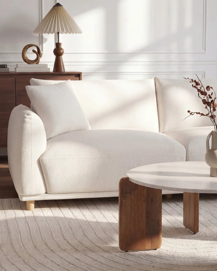 A cream-colored upholstered sofa in a bright, modern living room setting with a wooden side table, lamp, and neutral rug.