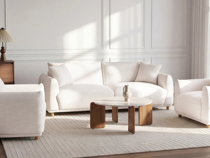 A bright, modern living room featuring a white three-seater sofa, a matching armchair, a round wooden coffee table, and a textured cream rug. The room has white wall paneling and is illuminated by soft, natural light coming from the side.