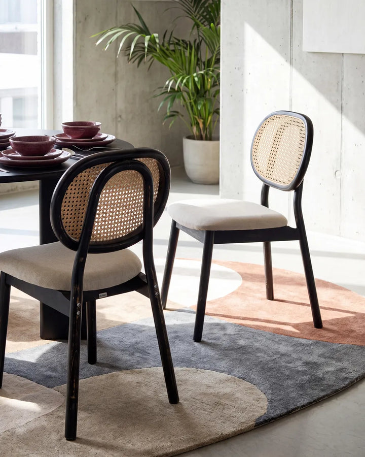 Two modern dining chairs with black frames and woven cane backrests, shown in a bright, contemporary room with concrete walls, a potted plant, and a patterned rug.