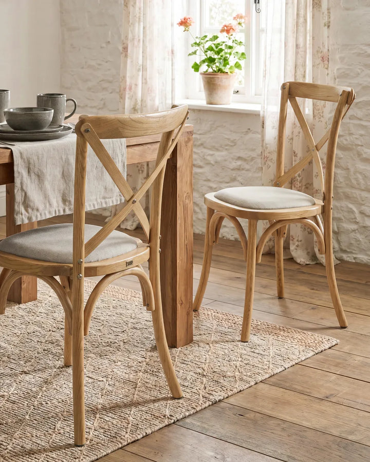 Two wooden dining chairs with light beige upholstered seats, shown in a bright, rustic dining room setting with a table, window, and potted plant.