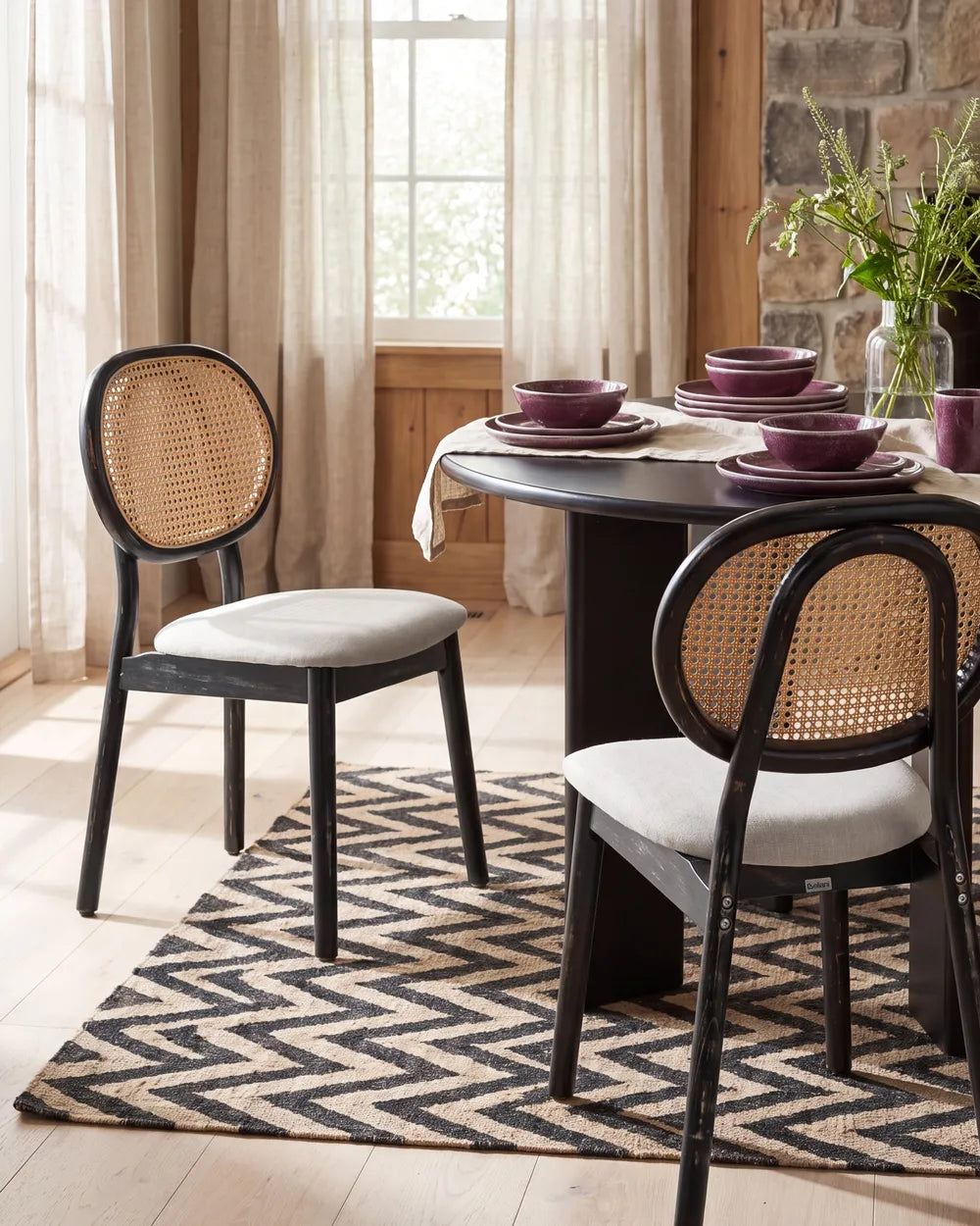 A dining room scene featuring two black dining chairs with woven cane backrests and light beige seat cushions, arranged around a dark round table on a black and beige chevron-patterned rug. The room has a stone wall, beige curtains, and a window in the background.