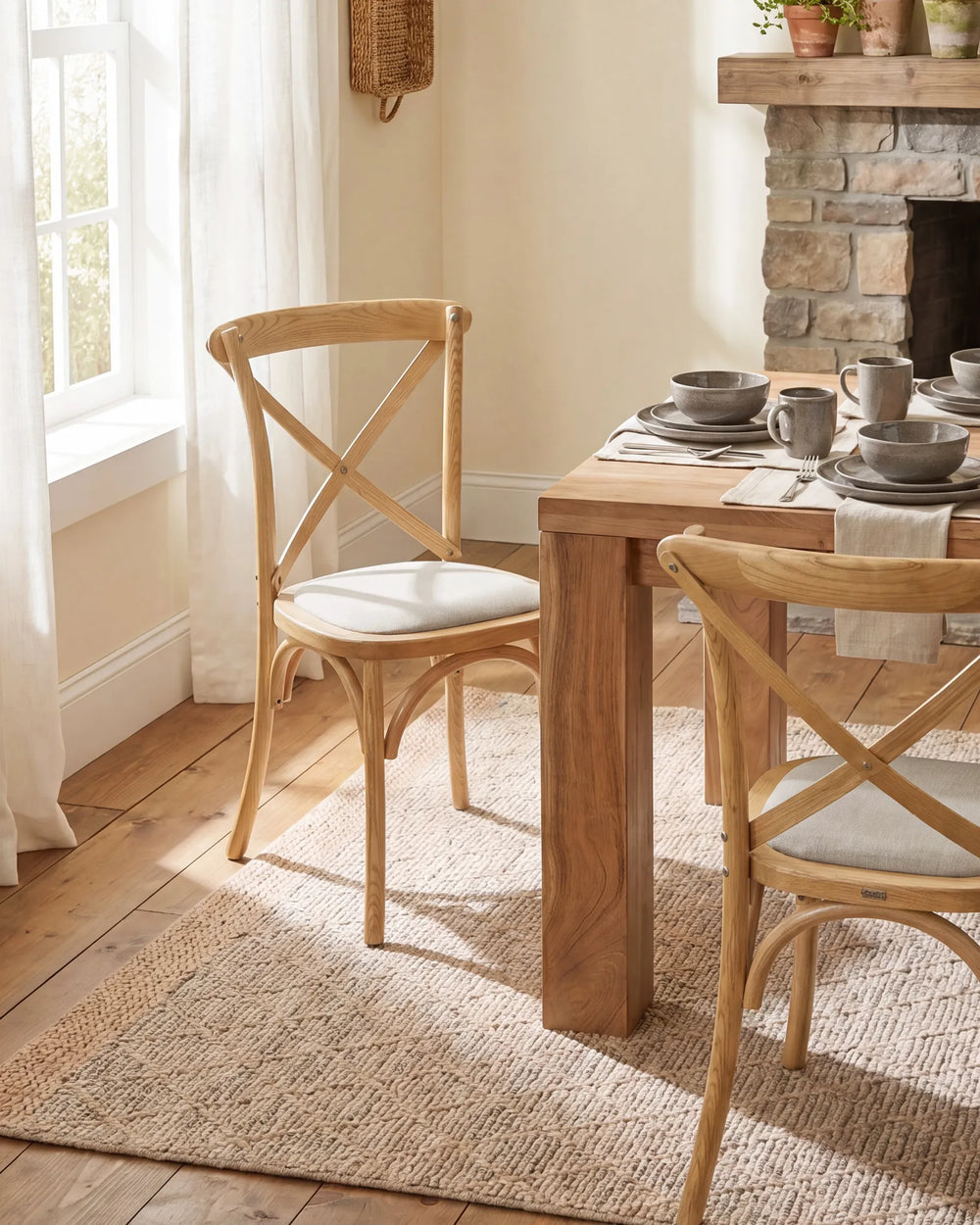 A dining room scene showing a light natural wood dining table with two matching wooden chairs that have cream upholstered seats. The table is set with gray ceramic mugs and plates, placed on a woven jute rug in front of a stone fireplace and next to a window with white curtains.