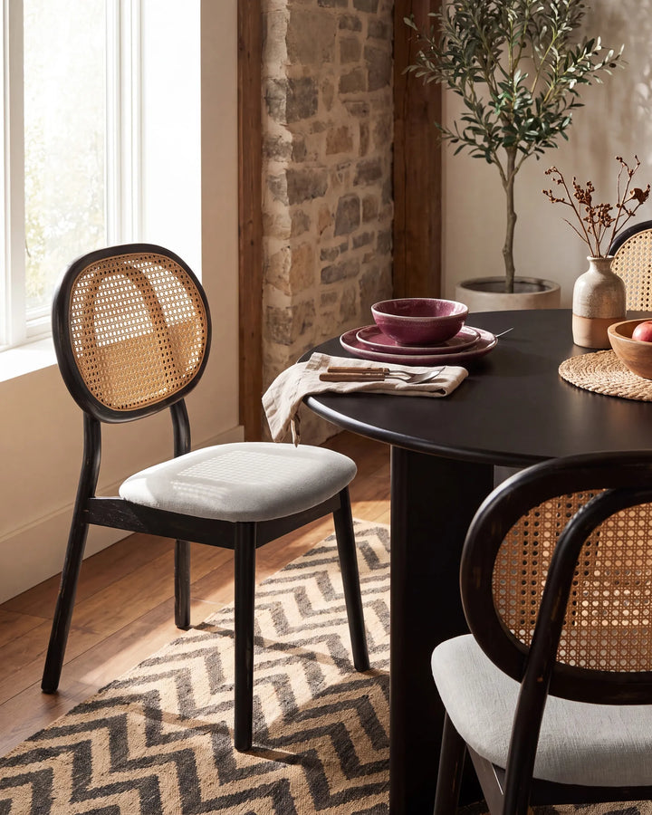 A dining room scene featuring a dark wood dining table with woven cane back chairs and a patterned rug, set in a warm, rustic interior with a stone wall and window.