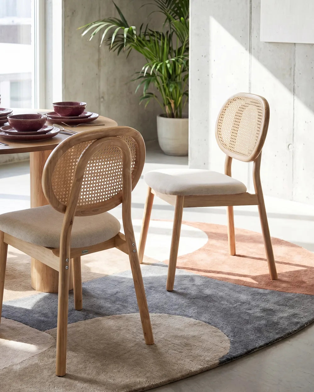 Two wooden dining chairs with woven cane backs and light beige upholstered seats, shown in a bright modern dining room setting with a table, rug, and potted plant.