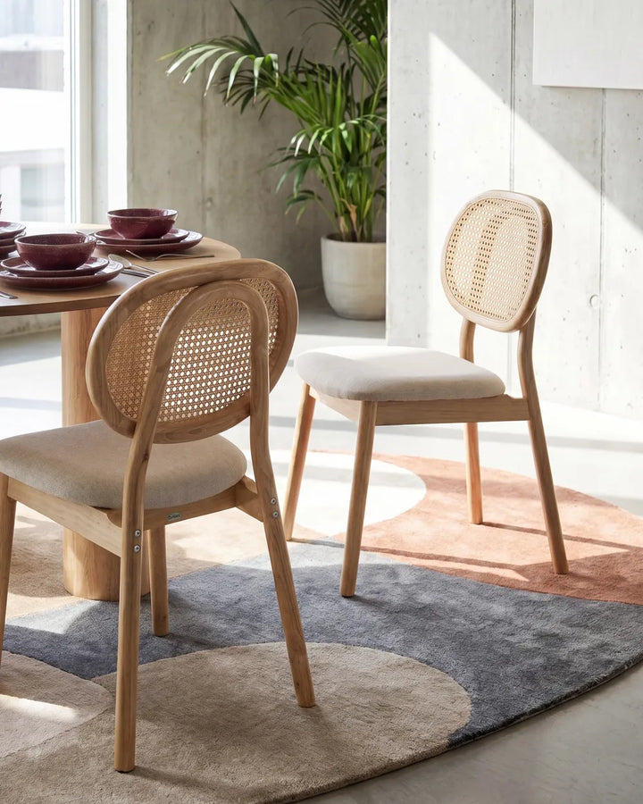 Two wooden dining chairs with woven cane backs and light beige upholstered seats, shown in a bright modern dining room setting with a table, rug, and potted plant.
