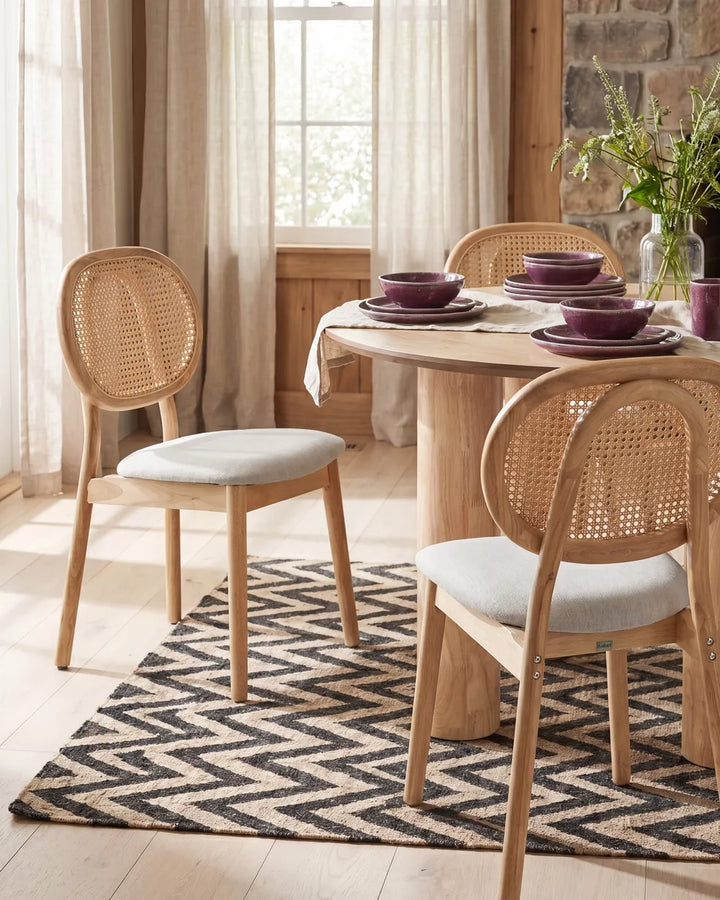 A dining room scene with a round wooden table and three woven cane chairs with light gray upholstered seats, set on a black and beige patterned rug near a window with sheer curtains.