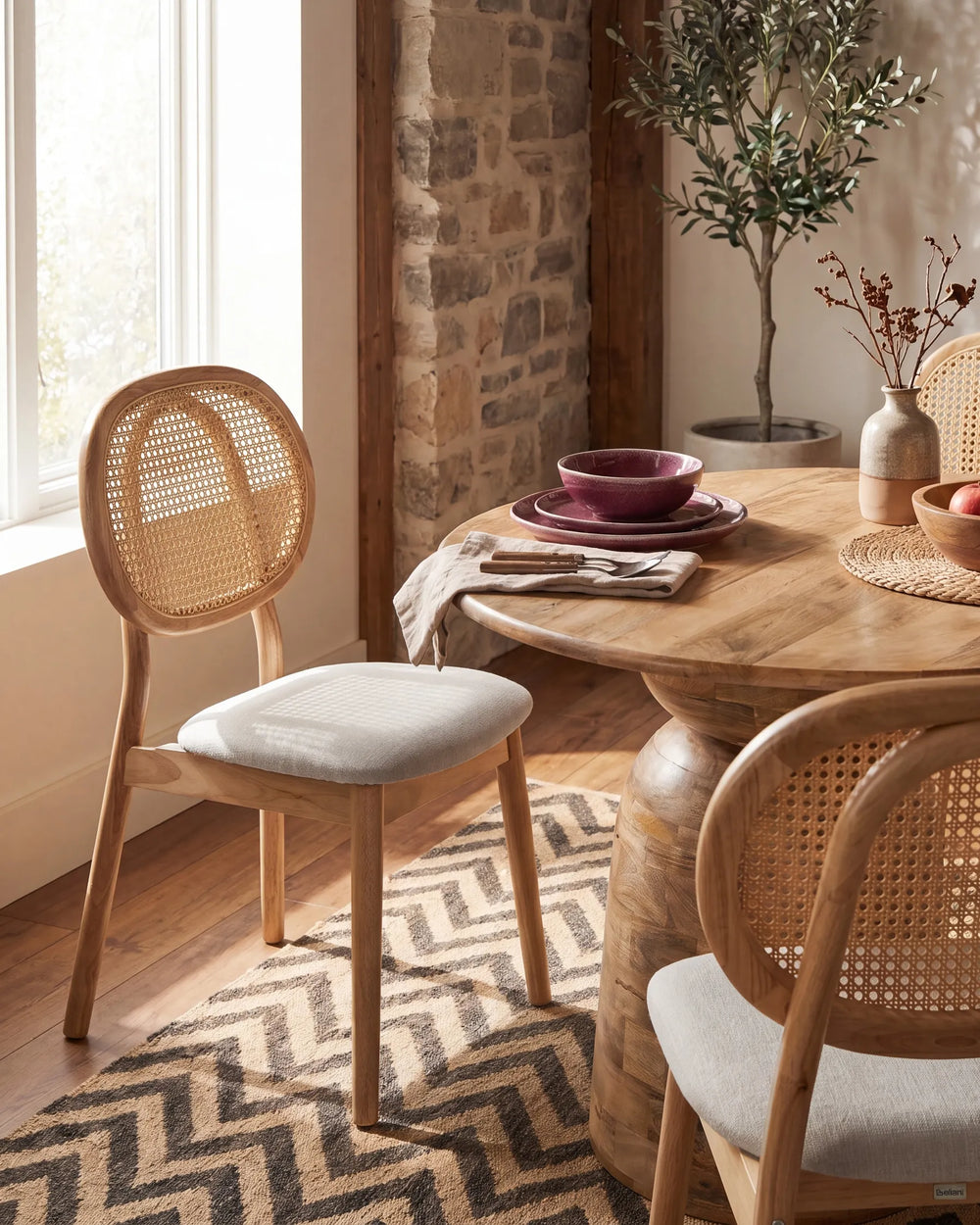 A warm, rustic dining room scene featuring a round wooden table, woven cane chairs with light beige cushions, a patterned rug, and a stone wall in the background.