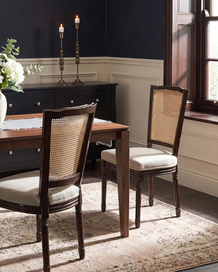 A dining room scene showing two wooden dining chairs with woven cane backs and light beige upholstered seats, placed around a wooden table on a patterned rug in a styled interior.