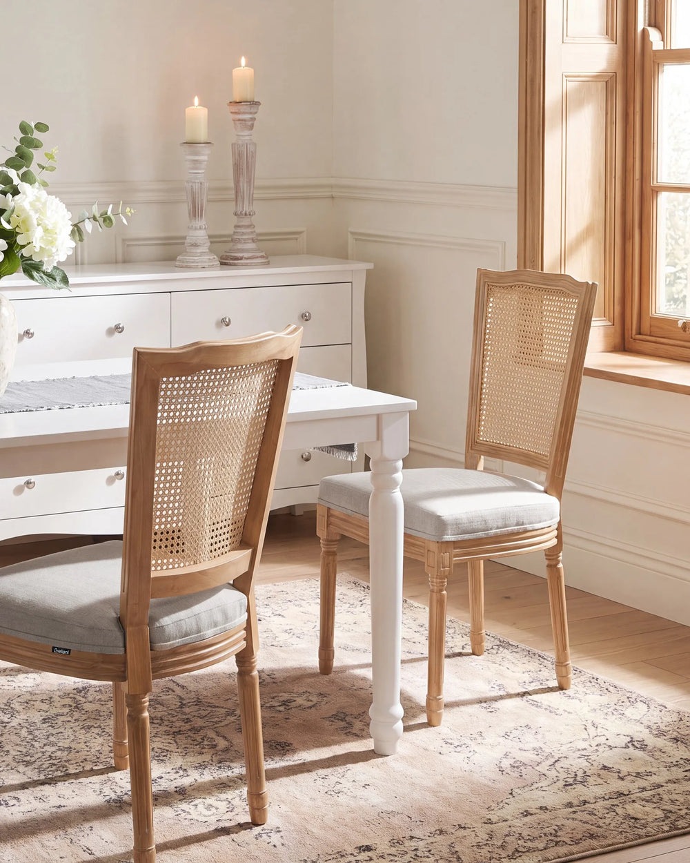 A dining room scene with two light wood chairs featuring woven cane backs and light gray upholstered seats, placed around a white table on a patterned rug in a bright, neutral interior.