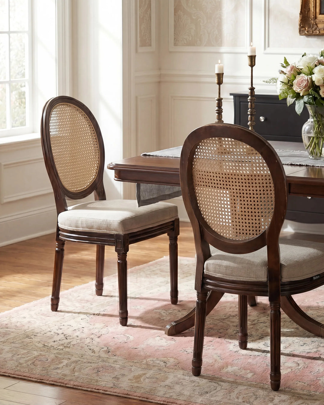 Two dark wood dining chairs with oval cane-back frames and light beige upholstered seats, shown in a styled dining room setting with a wooden table, rug, and flowers.