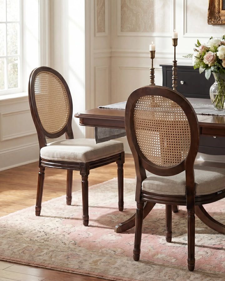 Two dark wood dining chairs with oval cane-back frames and light beige upholstered seats, shown in a styled dining room setting with a wooden table, rug, and flowers.