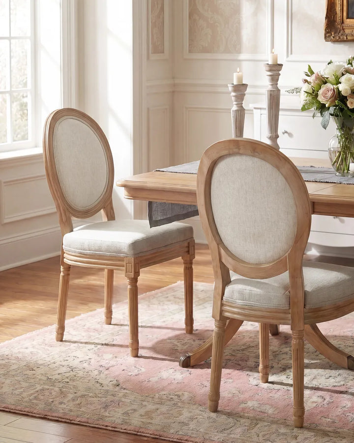 Two round-back dining chairs with light wood frames and cream upholstered seats, shown in a bright dining room beside a wooden table and floral centerpiece.