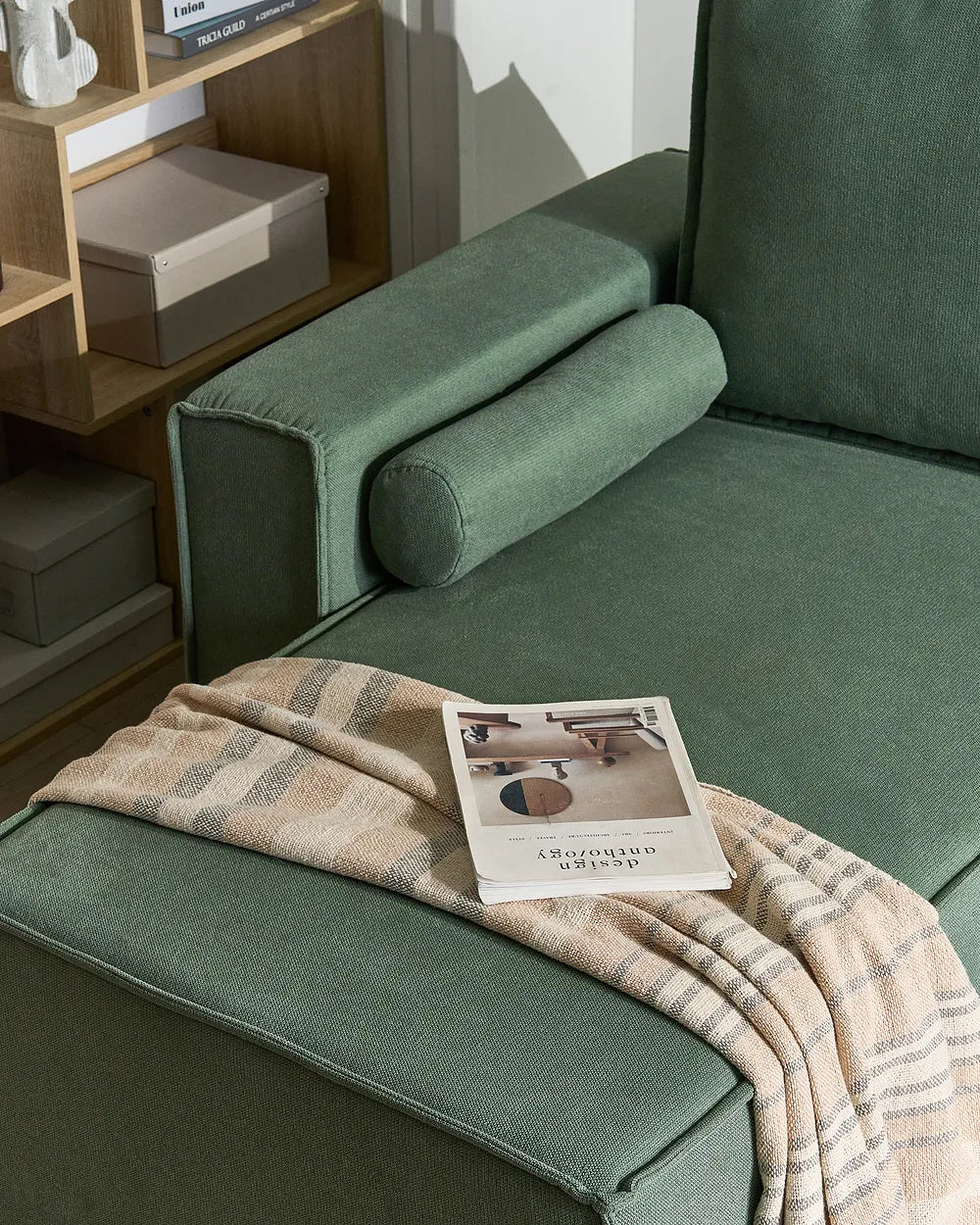 A green upholstered lounge chair or chaise in a styled interior, with a beige plaid throw blanket draped across the seat and a magazine resting on it. A wooden shelf with boxes is visible in the background.
