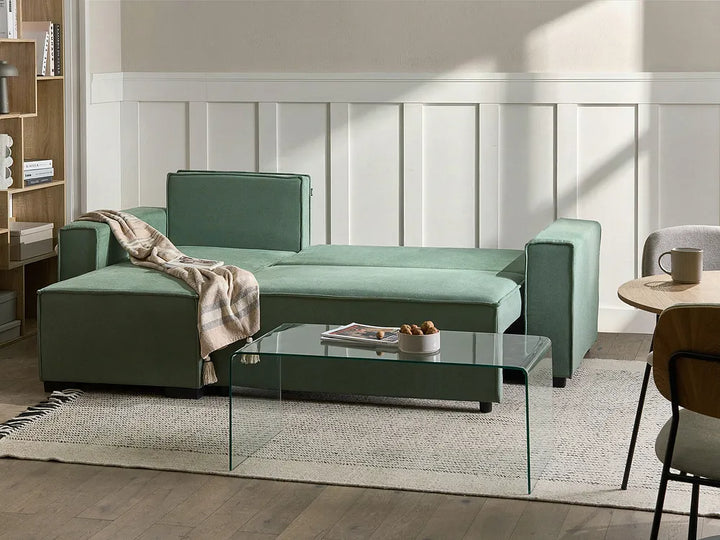 A sage green sectional sofa in a modern living room setting, styled with a beige throw blanket, a glass coffee table, and a light rug.