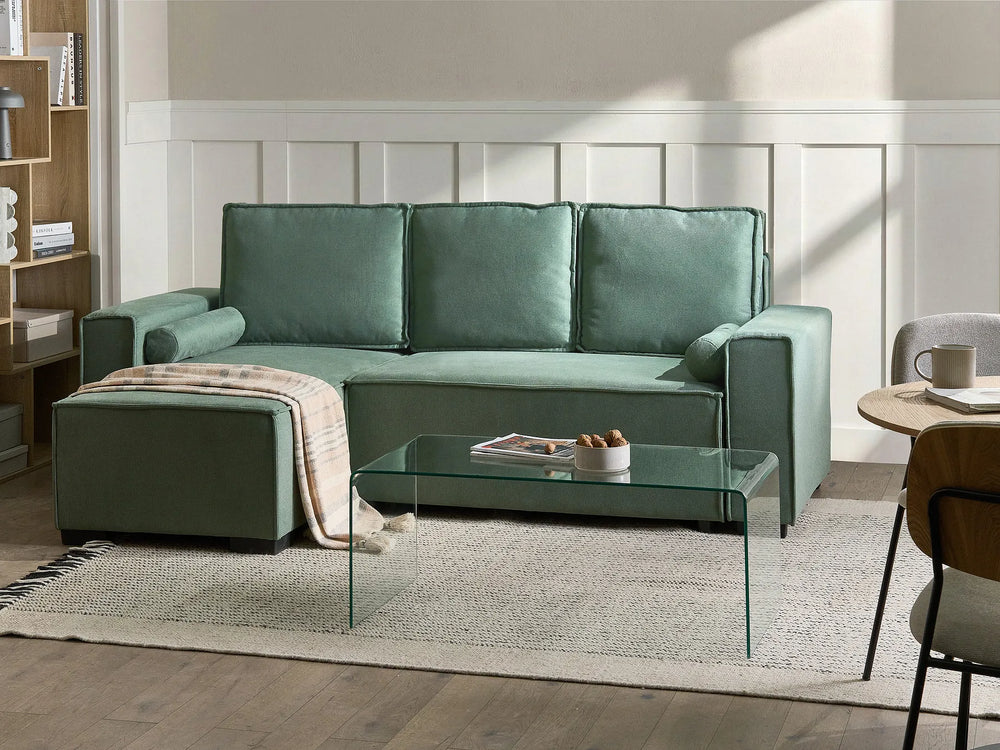 A sage green modular sectional sofa with a chaise lounge, styled in a bright, modern living room with a glass coffee table, textured rug, and wood paneling.