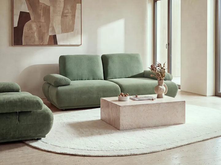 A modern living room featuring a sage green velvet sofa and matching armchair arranged around a light stone coffee table on a round white rug. The room has warm neutral walls, a large abstract painting, and tall windows with soft sunlight.