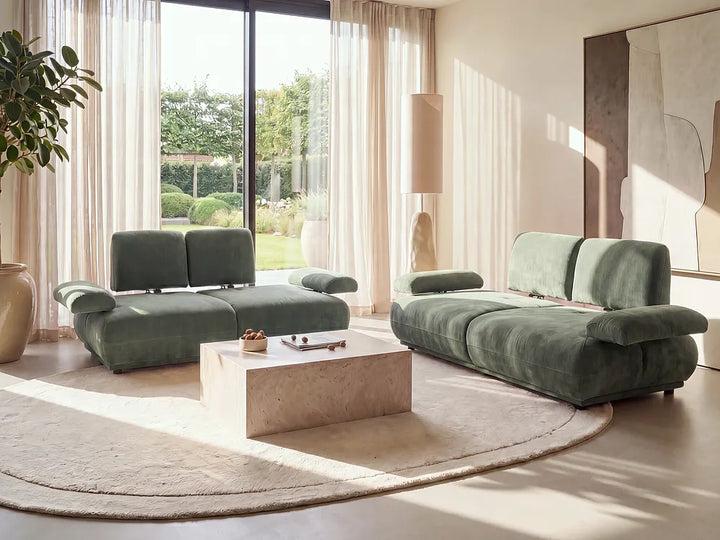 A modern living room featuring a sage green velvet sectional sofa and a matching chaise lounge set on a round beige rug, with a light stone coffee table in the center.