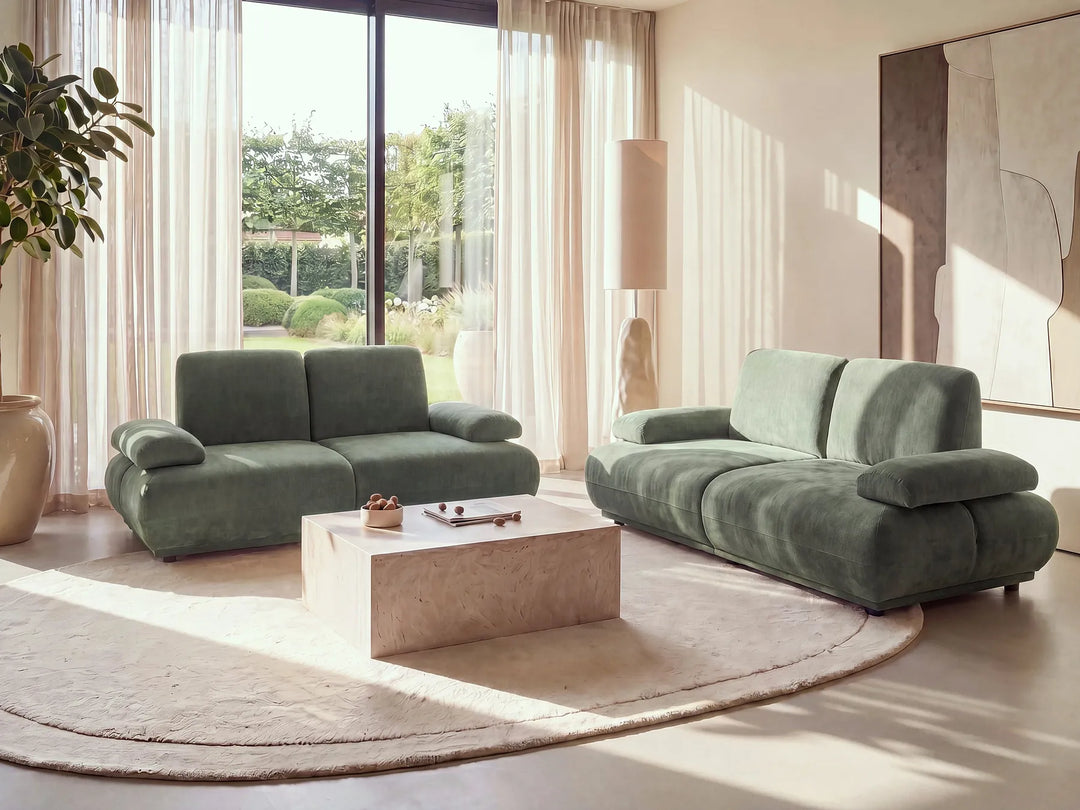 A modern living room featuring a sage green modular sofa set, including a two-seater loveseat and a three-seater sofa, arranged around a light stone coffee table on a round beige rug.