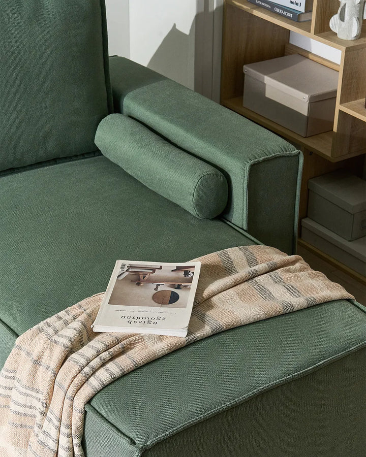 A green upholstered armchair in a styled interior, with a beige plaid throw blanket draped over the seat and a magazine resting on it. A wooden shelf with storage boxes is visible in the background.