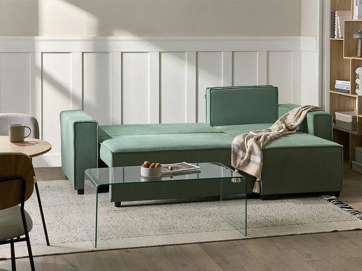 A modern sage green sectional sofa in a bright, contemporary living room setting with a glass coffee table and a beige throw blanket.