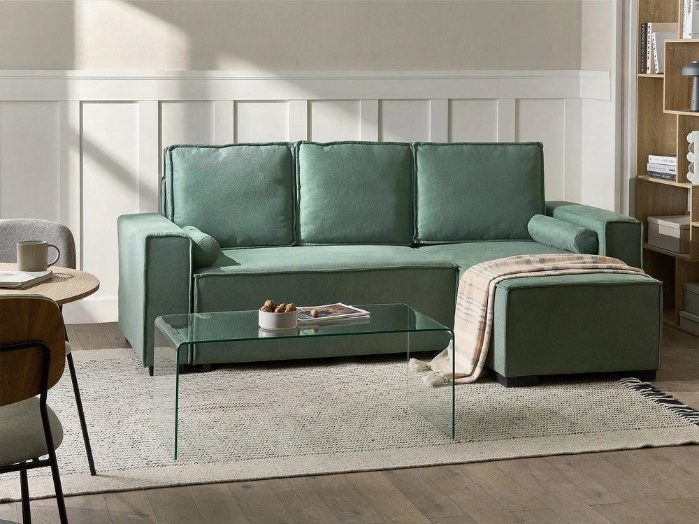 A sage green velvet sectional sofa with a chaise lounge, styled in a bright, modern living room with a glass coffee table, a light-colored rug, and a beige throw blanket.