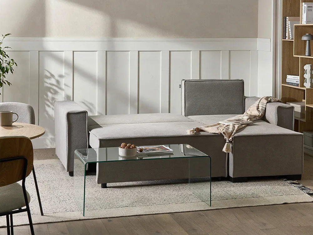 A modern gray fabric sectional sofa with a chaise lounge, styled in a bright, contemporary living room with a glass coffee table, textured rug, and white paneled wall.