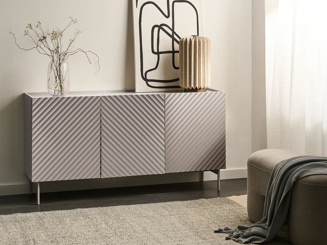 A modern sideboard cabinet with three doors featuring a diagonal ribbed texture, styled in a minimalist room with a vase of branches, a pleated gold lamp, and abstract wall art.