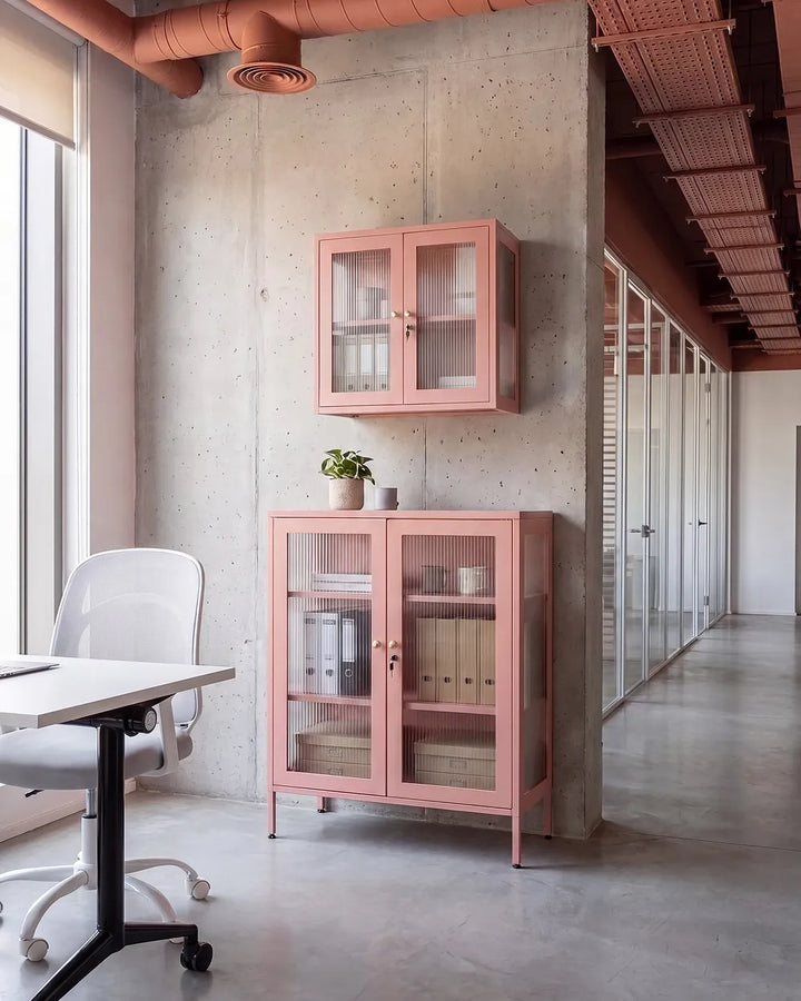 A pink metal office cabinet with glass doors stands against a concrete wall in a modern office space, with a matching smaller wall-mounted cabinet above it. A white desk chair and desk are visible at left, and a glass-walled corridor runs along the right.