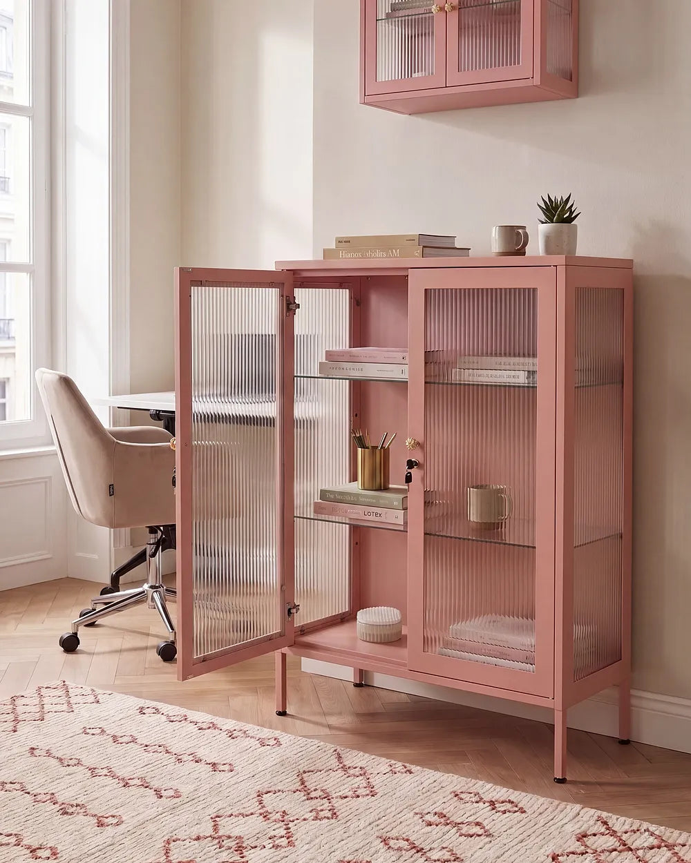 A dusty pink wooden cabinet with ribbed glass doors stands in a bright home office next to a desk and chair. The cabinet has three shelves and is styled with books, a mug, and a small plant.