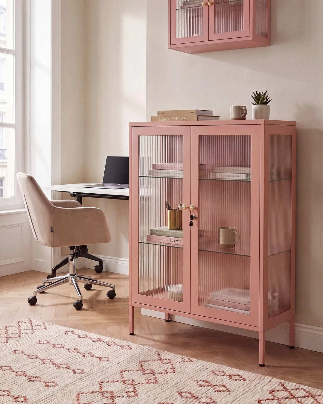 A dusty pink glass-front cabinet with ribbed doors stands in a bright home office next to a desk and chair. A matching pink wall cabinet hangs above, and a patterned rug covers the wood floor.