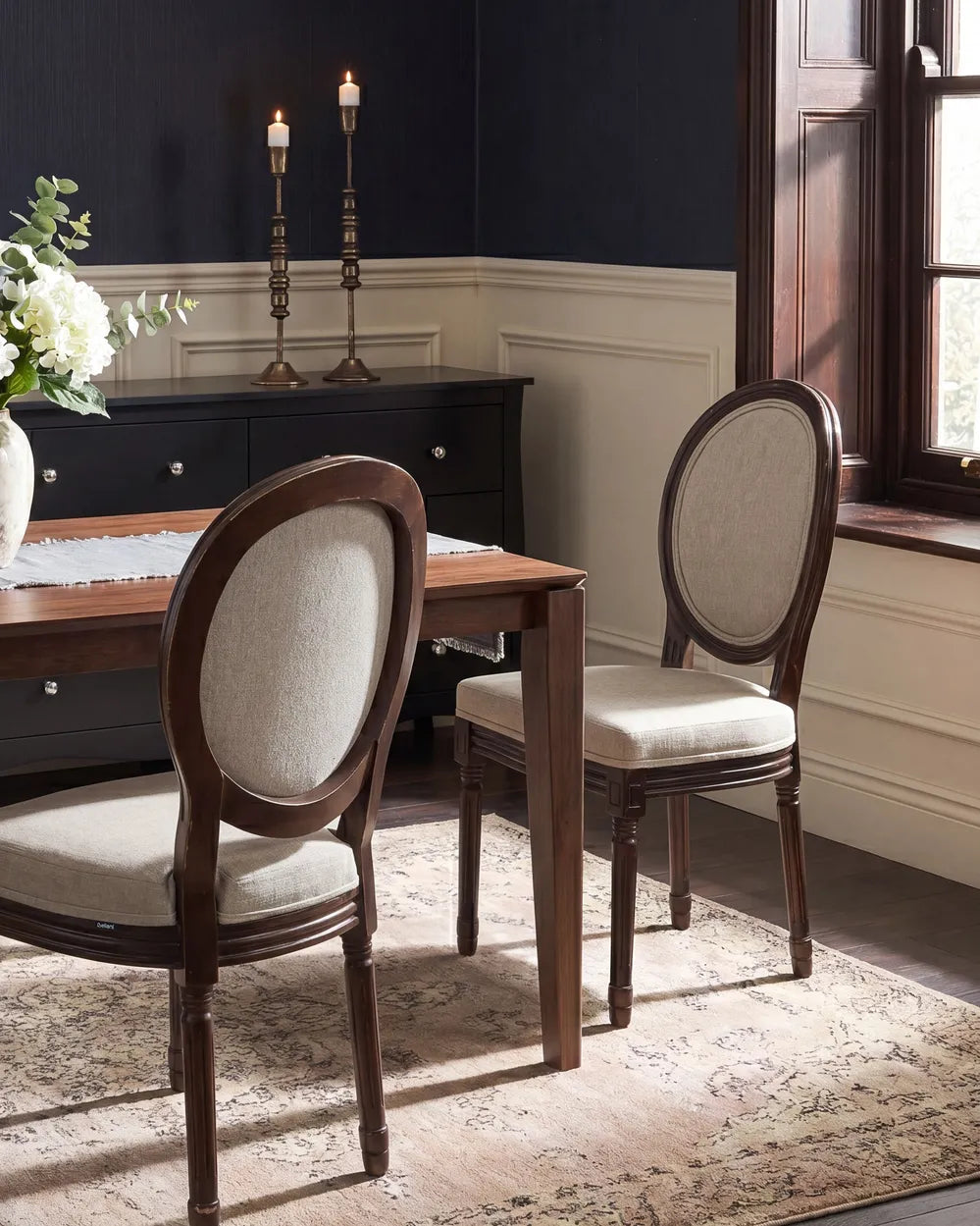 A dining room scene showing two upholstered wooden chairs with oval backs and light beige fabric seats, placed around a wooden dining table on a patterned rug. A dark wood sideboard, navy wall, white wainscoting, and a window are visible in the room.
