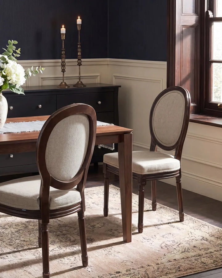 A dining room scene showing two upholstered wooden chairs with oval backs and light beige fabric seats, placed around a wooden dining table on a patterned rug. A dark wood sideboard, navy wall, white wainscoting, and a window are visible in the room.