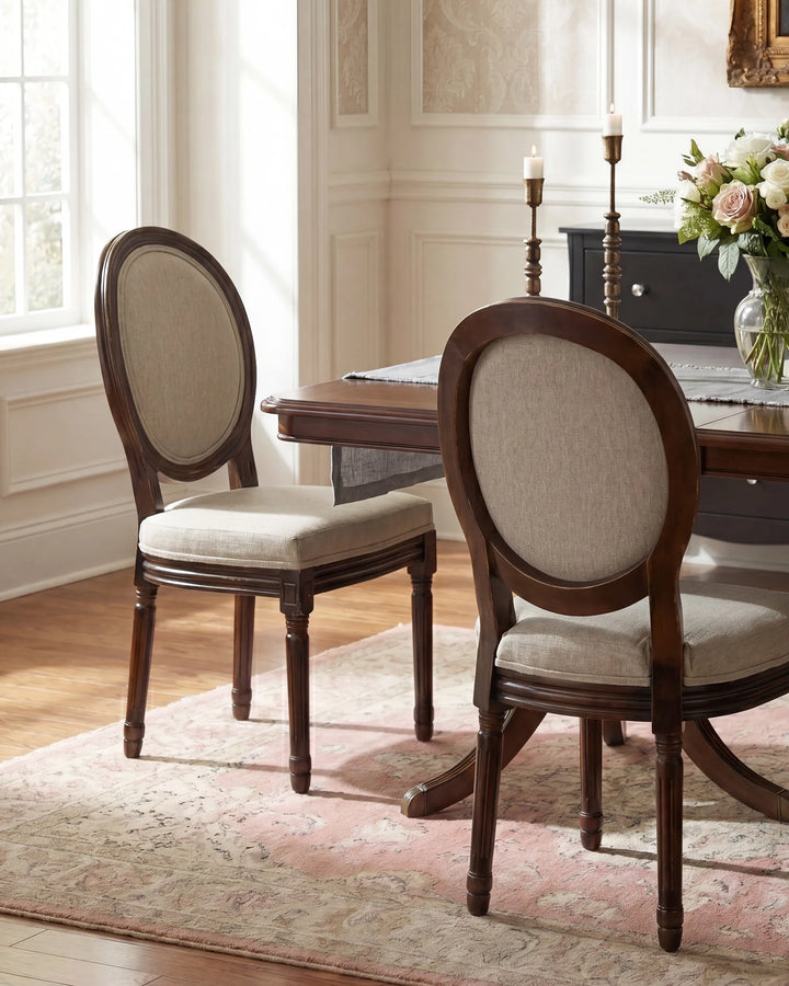 Two dark wood dining chairs with oval backrests and light beige upholstered seats, shown in a bright dining room beside a wooden table and floral centerpiece.