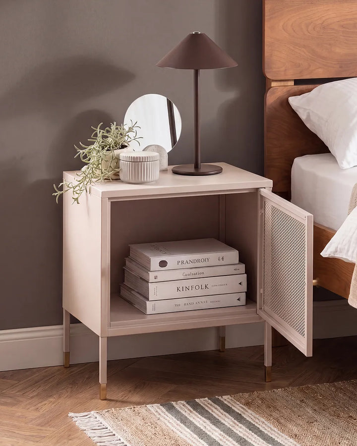 A light blush pink nightstand with an open door, styled in a bedroom next to a wooden bed frame. The nightstand features a single open shelf containing books, a small round mirror, a textured vase with greenery, and a dark bronze table lamp.