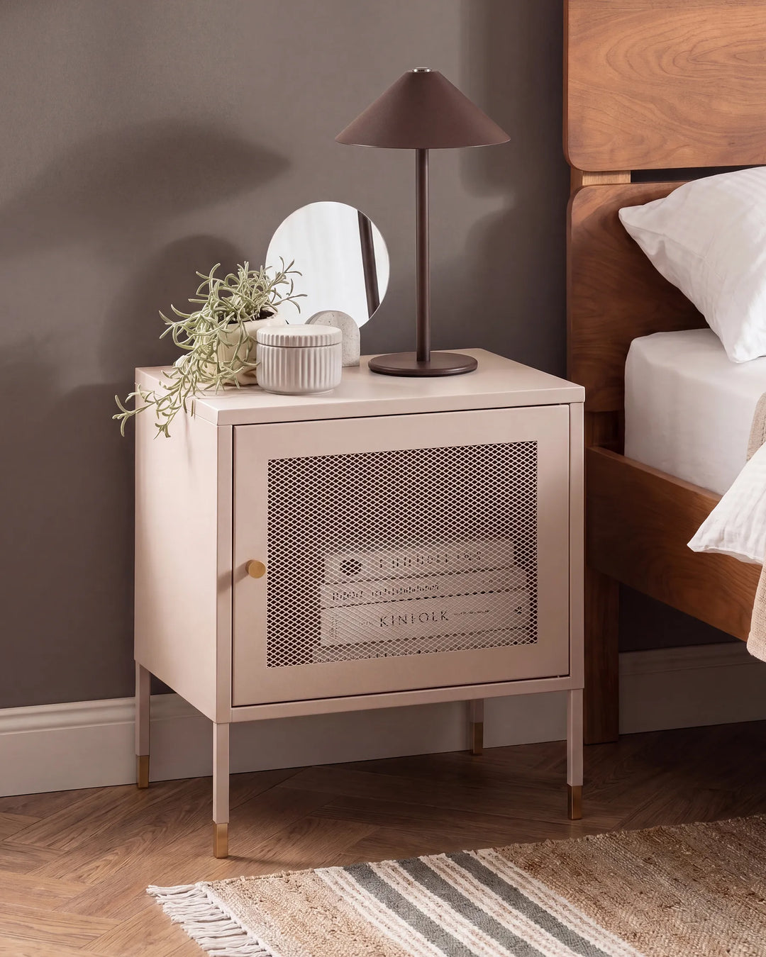 A light blush pink nightstand with a perforated metal door and gold legs, styled in a bedroom next to a wooden bed frame with a lamp and a small mirror on top.