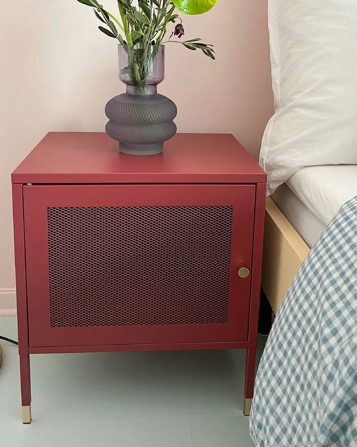 A red metal nightstand with a perforated front door and gold-tone leg caps, styled next to a bed with a gray ribbed glass vase on top.