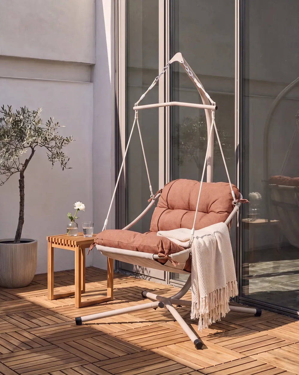 A hanging outdoor swing chair with a cushioned rust-brown seat and backrest, suspended by white ropes from a white metal frame. It is styled on a wooden deck next to a small wooden side table with a white throw blanket draped over it, in front of a glass sliding door.