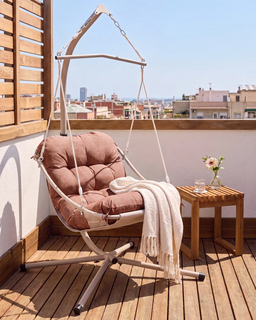 A cushioned hanging patio swing chair with a dusty rose cushion and cream rope, suspended from a metal frame on a wooden deck balcony. A cream throw blanket is draped over the seat, and a small wooden side table with a vase of flowers sits beside it.