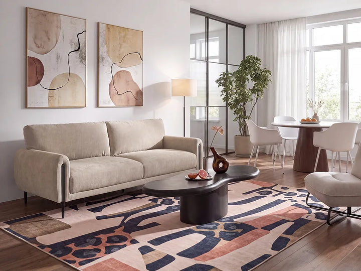 A modern living room featuring a light beige fabric sofa, a dark oval coffee table, a large abstract patterned area rug, and two framed abstract wall art prints on a white wall.