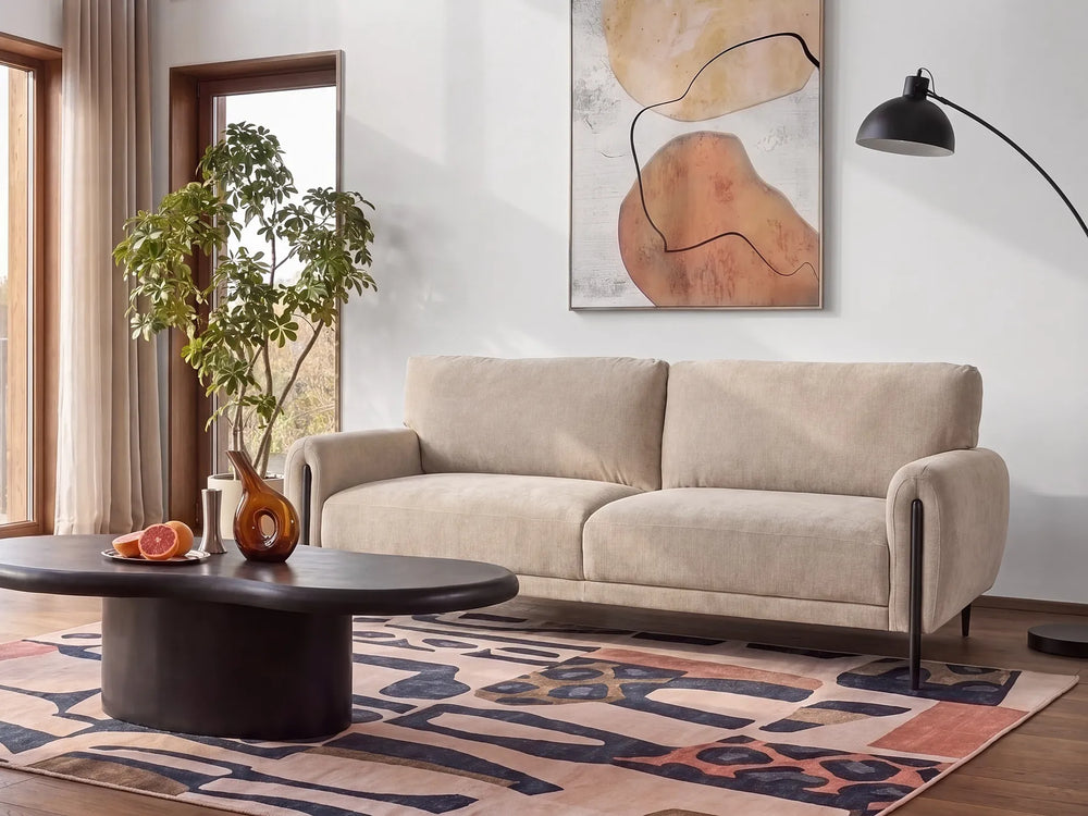 A modern beige two-seater sofa in a styled living room setting, positioned on a patterned rug next to a black oval coffee table and under a black floor lamp.