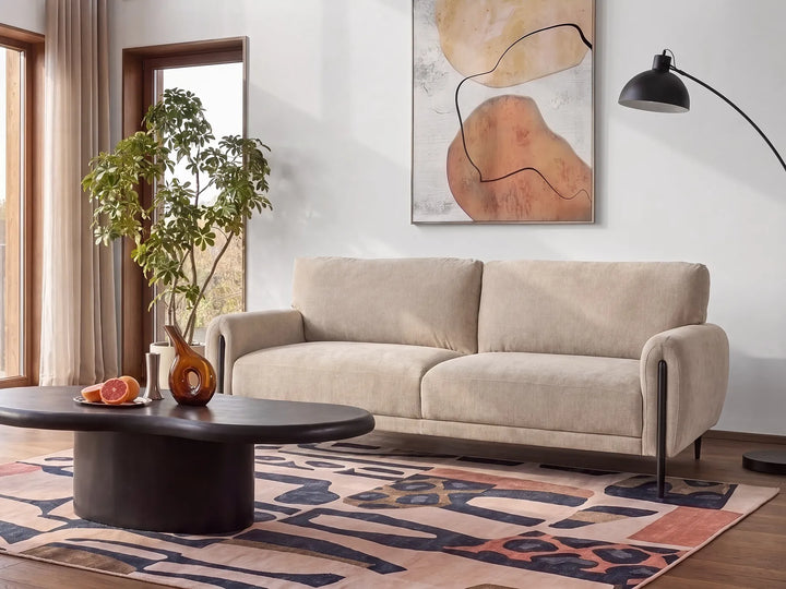 A modern beige two-seater sofa in a styled living room setting, positioned on a patterned rug next to a black oval coffee table and under a black floor lamp.