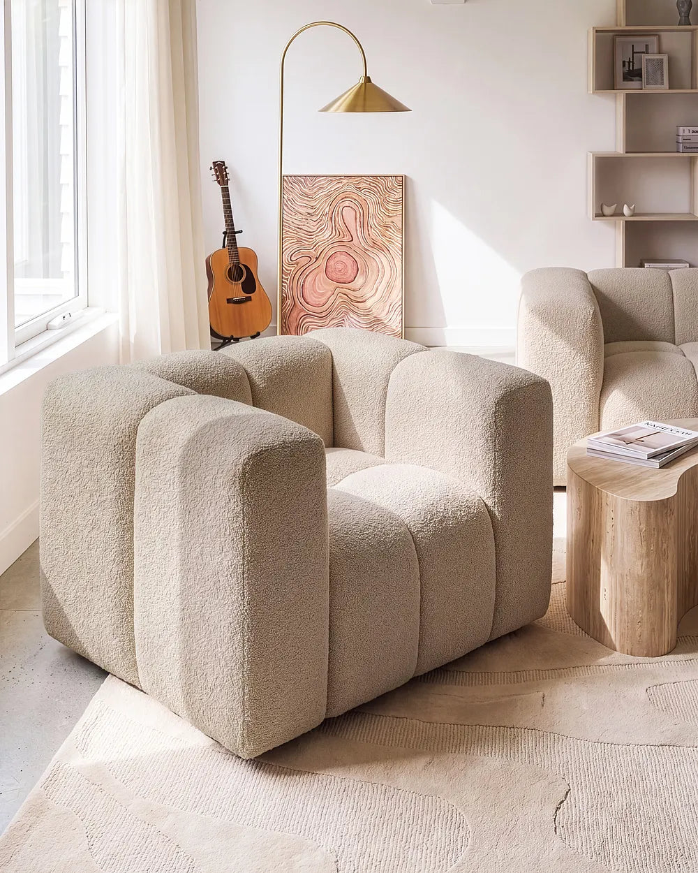 A modern beige bouclé armchair with a unique segmented, rounded backrest, styled in a bright, minimalist living room next to a wooden side table and a matching armchair.