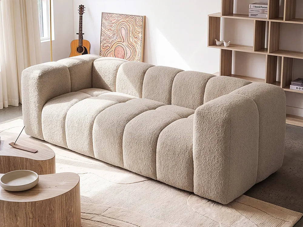 A modern beige bouclé sofa with a segmented, bubble-like design, styled in a bright, minimalist living room with wooden tables, a bookshelf, and an acoustic guitar in the background.